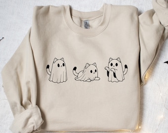 May include: Beige sweatshirt featuring three black and white cat ghost illustrations. The cats are in different poses, with one standing, one sitting, and one with its arms out. Halloween-themed apparel.