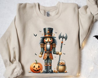 Puede incluir: Sudadera beige con un diseño de Halloween. El gráfico incluye un soldado cascanueces con un hacha, una calabaza tallada y un cubo de nueces. Siluetas de murciélagos negros están dispersas alrededor del diseño.