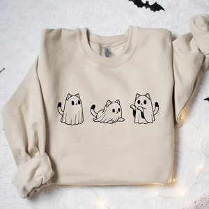 May include: Beige sweatshirt featuring three black and white cat ghost illustrations. The cats are in different poses, with one standing, one sitting, and one with its arms out. Halloween-themed apparel.