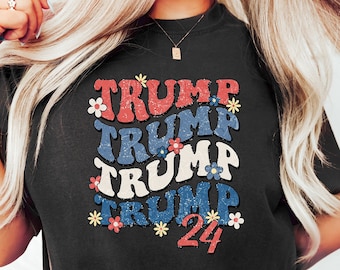 May include: A black t-shirt with a graphic design featuring the word "TRUMP" repeated four times in a retro font with red, white, and blue accents. The graphic also includes small white flowers. The number "24" is printed below the word "TRUMP".