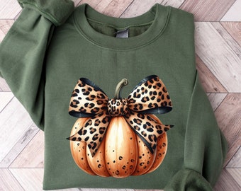 May include: Olive green sweatshirt featuring a watercolor pumpkin design. The pumpkin is orange with a leopard print bow. The sleeves are cuffed. The design is perfect for fall and autumn.