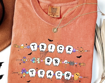 May include: A coral-colored Comfort Colors t-shirt with the words "TRICK OR TEACH" in a Halloween-themed design. The design includes skulls, candy, and other spooky elements. A gold necklace and a brown hat are also visible.