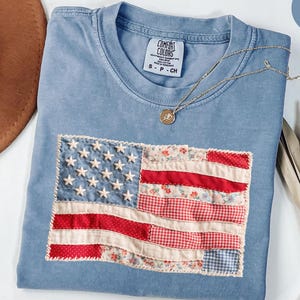 May include: A blue jean Comfort Colors t-shirt with a sewn-on American flag design. The flag features white stars and red and floral print stripes. A gold necklace with a pendant rests on the shirt.