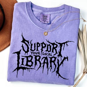 May include: A light purple short-sleeve t-shirt with a black graphic print that reads "SUPPORT YOUR LOCAL LIBRARY" in a jagged, stylized font. The shirt features a Comfort Colors brand label at the inner neckline and a crew neck collar.