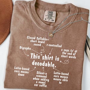 May include: A brown short-sleeved t-shirt with white text that reads: This shirt is decodable. Arrows point to linguistic notes including Closed Syllable, r-controlled, Digraphs, s says /z/, Latin-based roots, and silent-e rules. The shirt has a crew neck.