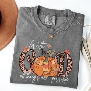 May include: A gray Comfort Colors t-shirt with the phrase "With God all things are possible." The design features an orange pumpkin wearing a scarf, leopard print letters, and fall leaves. A silver necklace is draped across the shirt.