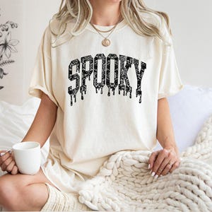 May include: Cream-colored t-shirt with the word "SPOOKY" in black, dripping letters. The shirt has a relaxed fit and is worn by someone sitting on a bed. A white mug is held in the hand.