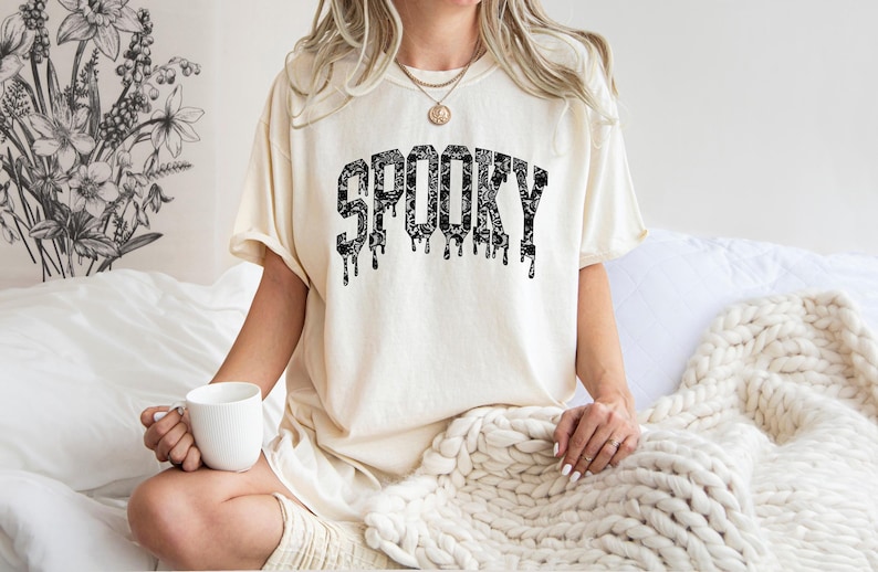 May include: Cream-colored t-shirt with the word "SPOOKY" in black, dripping letters. The shirt has a relaxed fit and is worn by someone sitting on a bed. A white mug is held in the hand.