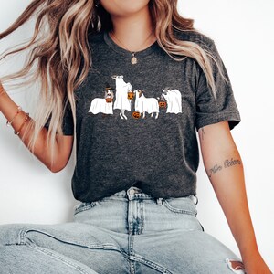May include: Dark gray t-shirt featuring a Halloween-themed design. The graphic depicts four dogs dressed as ghosts, each holding a jack-o'-lantern. One dog wears a witch's hat. A fun and festive design for dog lovers.