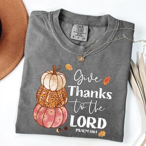 May include: A gray Comfort Colors t-shirt featuring a stacked pumpkin design in orange, white, and pink. The shirt has the text "Give Thanks to the LORD" and "PSALM 136:1" in white script. A gold necklace is draped across the shirt.