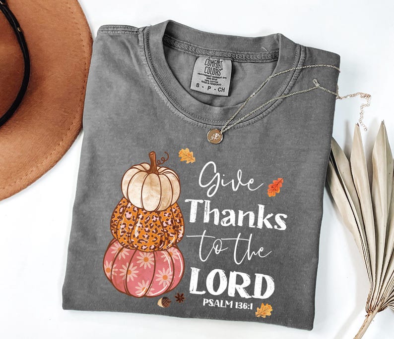 May include: A gray Comfort Colors t-shirt featuring a stacked pumpkin design in orange, white, and pink. The shirt has the text "Give Thanks to the LORD" and "PSALM 136:1" in white script. A gold necklace is draped across the shirt.