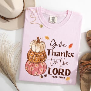 May include: Light pink t-shirt with a stacked pumpkin graphic and the text "Give Thanks to the LORD" with Psalm 136:1. The shirt is next to brown sandals, a beige hat, and decorative grass.
