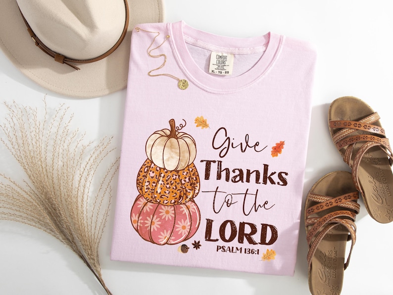 May include: Light pink t-shirt with a stacked pumpkin graphic and the text "Give Thanks to the LORD" with Psalm 136:1. The shirt is next to brown sandals, a beige hat, and decorative grass.