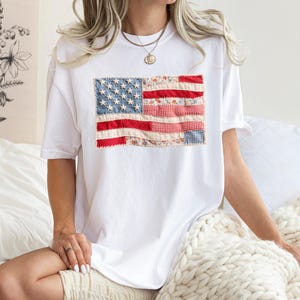 Peut inclure: T-shirt blanc avec un drapeau am&eacute;ricain en patchwork. Le drapeau utilise des tissus rouges, blancs et bleus avec des motifs floraux et vichy. Le t-shirt est de style d&eacute;contract&eacute; &agrave; manches courtes.