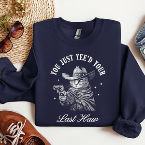 May include: Navy blue sweatshirt with a white graphic of a cat wearing a cowboy hat and holding a gun. The text "You just yee'd your last haw" is printed below the cat.