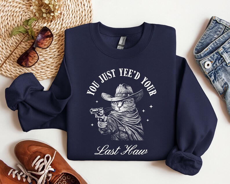 May include: Navy blue sweatshirt with a white graphic of a cat wearing a cowboy hat and holding a gun. The text "You just yee'd your last haw" is printed below the cat.