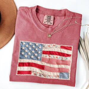Peut inclure: Un t-shirt rose fan&eacute; avec un drapeau am&eacute;ricain cousu. Le drapeau pr&eacute;sente des &eacute;toiles blanches sur un fond bleu et des rayures rouges et &agrave; motifs floraux. Un collier argent&eacute; avec un petit pendentif repose sur le t-shirt.