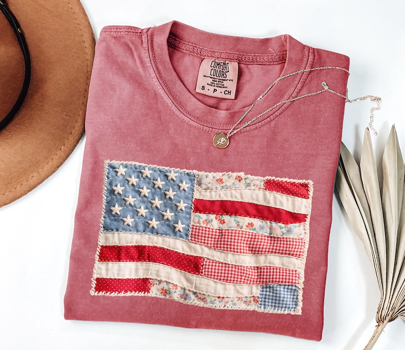 Peut inclure: Un t-shirt rose fan&eacute; avec un drapeau am&eacute;ricain cousu. Le drapeau pr&eacute;sente des &eacute;toiles blanches sur un fond bleu et des rayures rouges et &agrave; motifs floraux. Un collier argent&eacute; avec un petit pendentif repose sur le t-shirt.