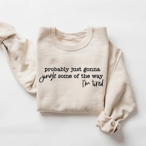 May include: A cream-colored sweatshirt with the text "probably just gonna jingle some of the way I'm tired" in black script. The sweatshirt is folded with the sleeves rolled up, and is displayed on a white background.