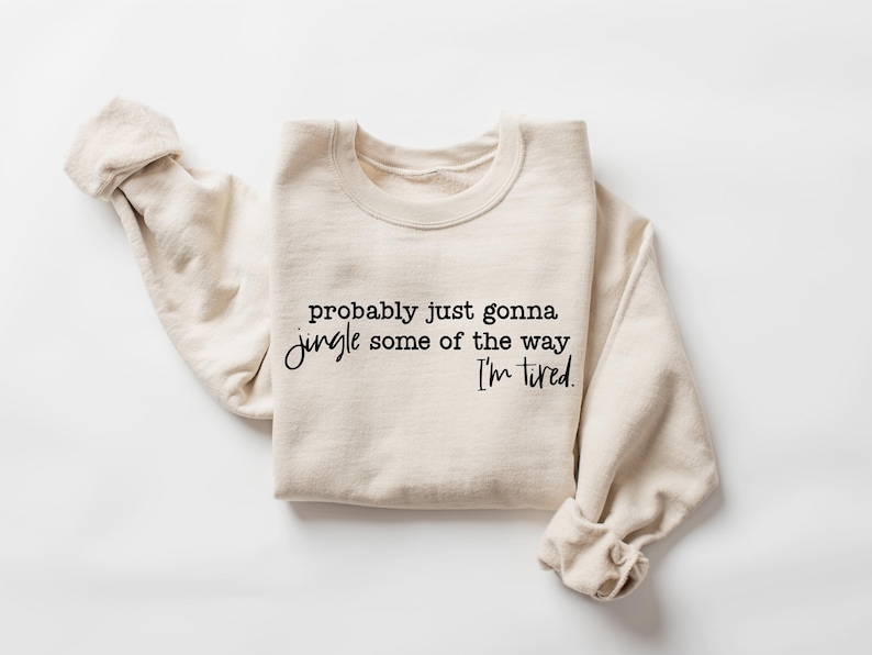 May include: A cream-colored sweatshirt with the text "probably just gonna jingle some of the way I'm tired" in black script. The sweatshirt is folded with the sleeves rolled up, and is displayed on a white background.