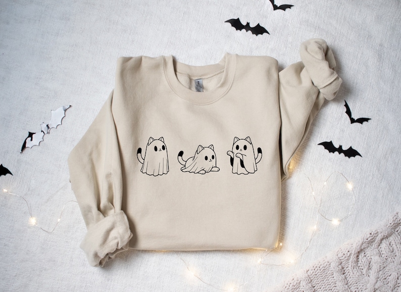 May include: Beige sweatshirt featuring three black and white cat ghost illustrations. The cats are in different poses, with one standing, one sitting, and one with its arms out. Halloween-themed apparel.