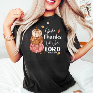 May include: Black t-shirt featuring a stacked pumpkin design with the text "Give Thanks to the LORD" and Psalm 136:1. The pumpkins are beige, leopard print, and pink with a floral pattern. Autumn leaves accent the design.
