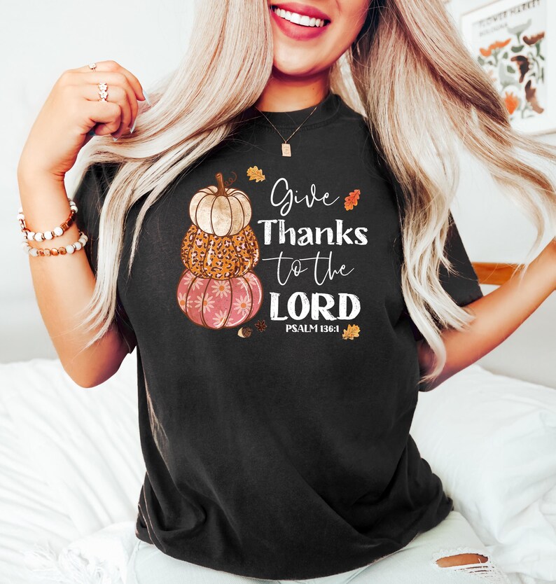May include: Black t-shirt featuring a stacked pumpkin design with the text "Give Thanks to the LORD" and Psalm 136:1. The pumpkins are beige, leopard print, and pink with a floral pattern. Autumn leaves accent the design.