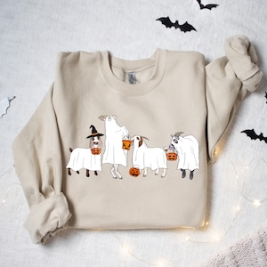 May include: Beige sweatshirt featuring a Halloween-themed graphic. The design depicts four goats dressed as ghosts, each holding a jack-o'-lantern. One goat wears a witch hat. Black bat cutouts and string lights decorate the background.