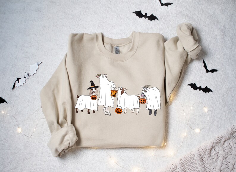 May include: Beige sweatshirt featuring a Halloween-themed graphic. The design depicts four goats dressed as ghosts, each holding a jack-o'-lantern. One goat wears a witch hat. Black bat cutouts and string lights decorate the background.