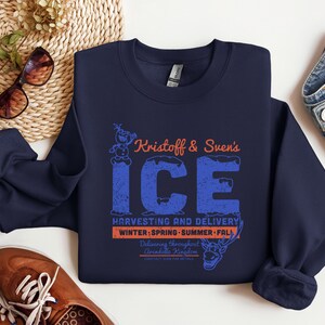 May include: Navy blue sweatshirt with a graphic featuring the text "Kristoff & Sven's ICE" in blue and orange. The design includes Olaf and Sven characters, with "Harvesting and Delivery" and seasonal references.