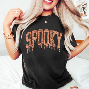 May include: Black t-shirt with the word "SPOOKY" in orange, dripping letters. The letters are filled with a pattern of bats and other Halloween-themed designs. The shirt is a comfortable, casual style.