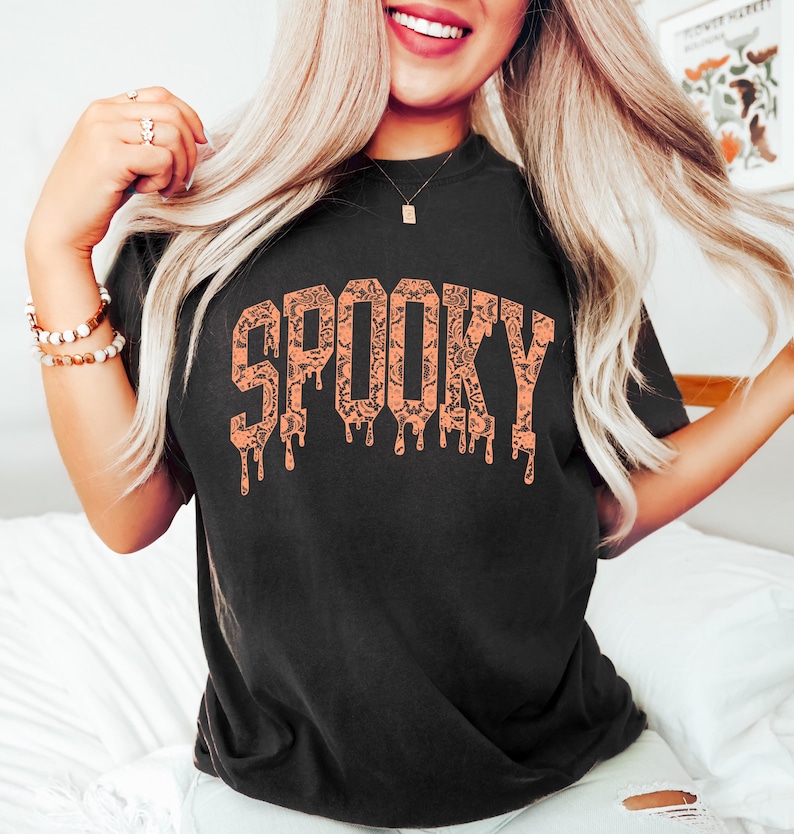 May include: Black t-shirt with the word "SPOOKY" in orange, dripping letters. The letters are filled with a pattern of bats and other Halloween-themed designs. The shirt is a comfortable, casual style.