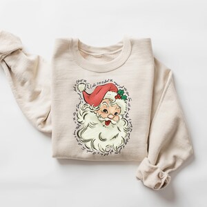 Pu&ograve; includere: Felpa girocollo beige con grafica vintage di Babbo Natale. L'illustrazione di Babbo Natale ha un cappello rosso, barba bianca e accenti di agrifoglio. Il testo circonda l'immagine. Un top comodo a maniche lunghe per le festivit&agrave;.