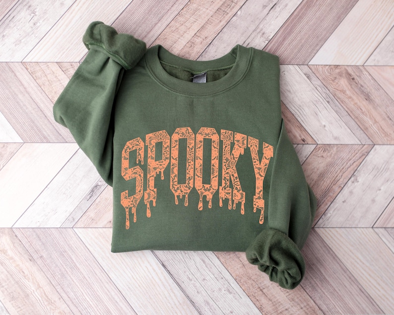 May include: An olive green sweatshirt with the word "SPOOKY" printed in orange. The letters have a dripping effect and a patterned design. The sweatshirt is folded on a light wood-grain surface.