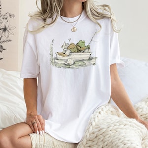 May include: A white short-sleeve t-shirt featuring a graphic illustration of two green frogs sitting in a small boat. One frog is reading a book while the other holds a fishing rod. The design includes water, a lily pad, and a dragonfly.