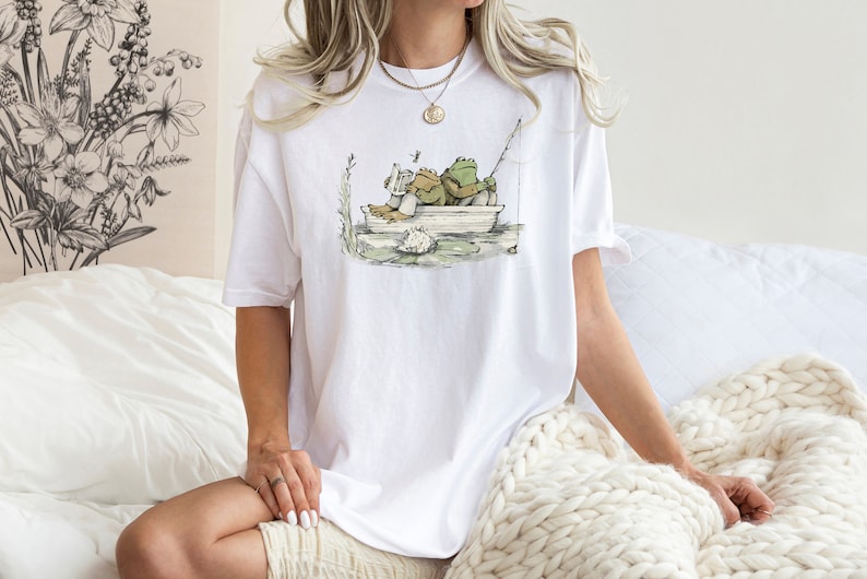 May include: A white short-sleeve t-shirt featuring a graphic illustration of two green frogs sitting in a small boat. One frog is reading a book while the other holds a fishing rod. The design includes water, a lily pad, and a dragonfly.