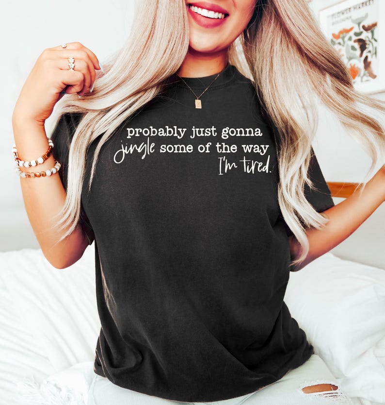 May include: Black t-shirt with the text "probably just gonna jingle some of the way, I'm tired" in white script. The shirt is a crew neck and appears to be made of a soft material. The shirt is worn by a person.