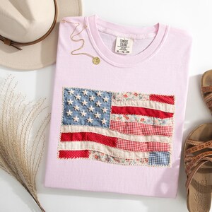 Peut inclure: Un t-shirt rose clair avec un drapeau am&eacute;ricain en patchwork. Le drapeau utilise des tissus rouges, blancs et bleus. Un collier dor&eacute; et des sandales marron sont &eacute;galement visibles. Un chapeau beige et de l'herbe s&eacute;ch&eacute;e compl&egrave;tent l'ensemble.