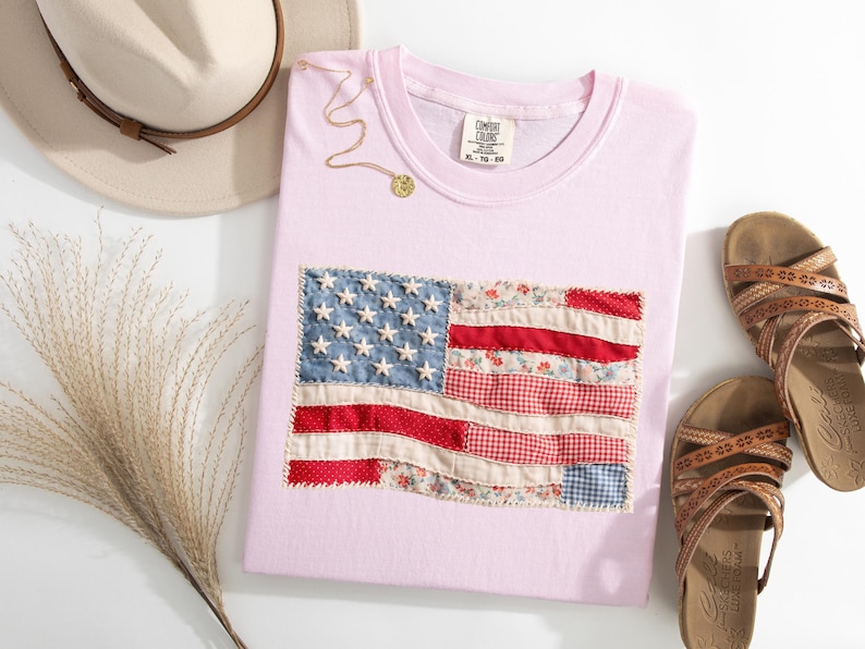 Peut inclure: Un t-shirt rose clair avec un drapeau am&eacute;ricain en patchwork. Le drapeau utilise des tissus rouges, blancs et bleus. Un collier dor&eacute; et des sandales marron sont &eacute;galement visibles. Un chapeau beige et de l'herbe s&eacute;ch&eacute;e compl&egrave;tent l'ensemble.
