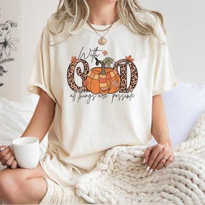 May include: Cream-colored t-shirt with a fall-themed graphic. The design features the words "With God all things are possible" with a pumpkin and leopard print letters. The shirt is a comfortable, casual style.