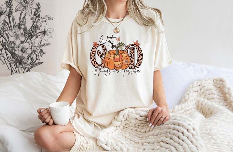 May include: Cream-colored t-shirt with a fall-themed graphic. The design features the words "With God all things are possible" with a pumpkin and leopard print letters. The shirt is a comfortable, casual style.