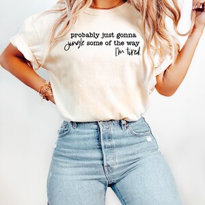 May include: Cream-colored t-shirt with the text "probably just gonna jingle some of the way I'm tired" in black script. The shirt is paired with light blue denim jeans.
