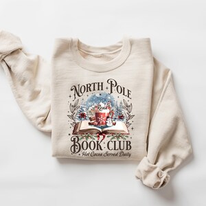May include: Cream-colored sweatshirt with a graphic that reads "North Pole Book Club" with a winter scene, a mug of hot cocoa, and an open book. The text "Hot Cocoa Served Daily" is also included. The sweatshirt is folded.