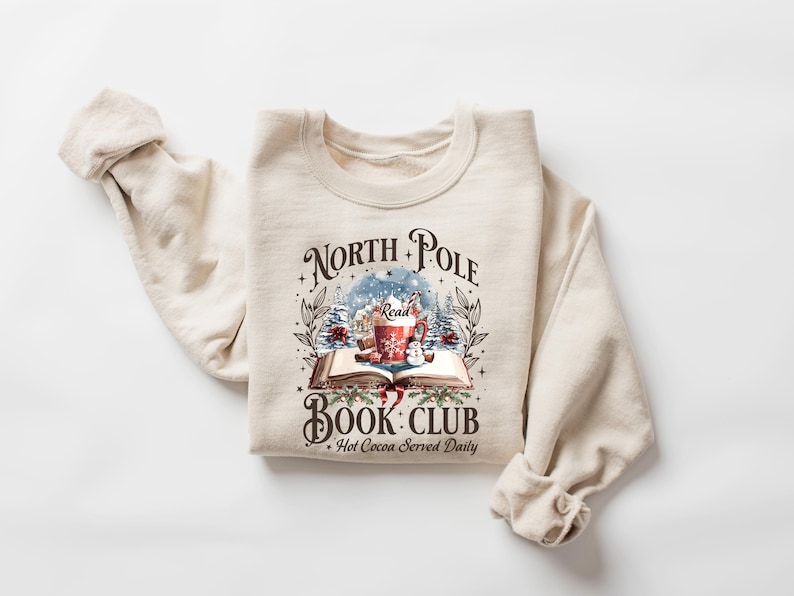 May include: Cream-colored sweatshirt with a graphic that reads "North Pole Book Club" with a winter scene, a mug of hot cocoa, and an open book. The text "Hot Cocoa Served Daily" is also included. The sweatshirt is folded.