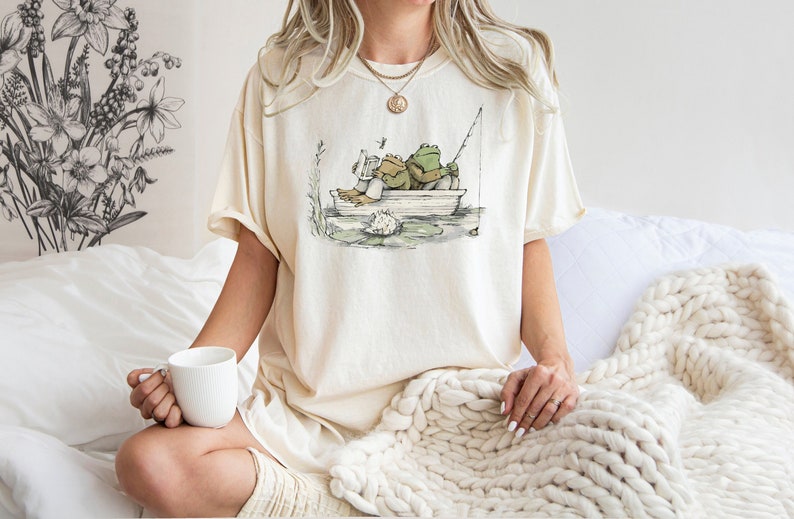 May include: A cream-colored t-shirt featuring a graphic illustration of two frogs sitting in a small boat on water. One frog is reading a book while the other holds a fishing rod. The design includes a lily pad and a small dragonfly in the air.