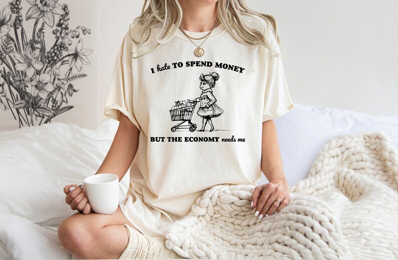 May include: A white t-shirt with the text "I hate to spend money but the economy needs me" and a black and white illustration of a woman pushing a shopping cart.