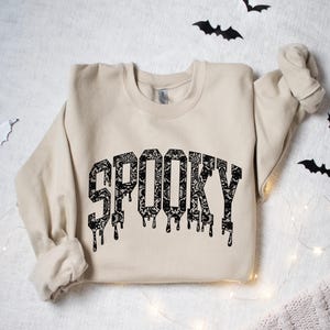 May include: A beige sweatshirt with the word "SPOOKY" in a black, dripping font. The letters are filled with a floral lace pattern. Black bat cutouts and string lights decorate the background, creating a Halloween theme.