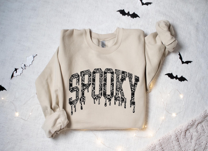May include: A beige sweatshirt with the word "SPOOKY" in a black, dripping font. The letters are filled with a floral lace pattern. Black bat cutouts and string lights decorate the background, creating a Halloween theme.