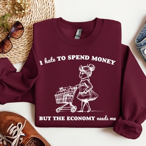 May include: Burgundy sweatshirt with white text that reads "I hate to spend money but the economy needs me". A cartoon illustration of a girl pushing a shopping cart is above the text.