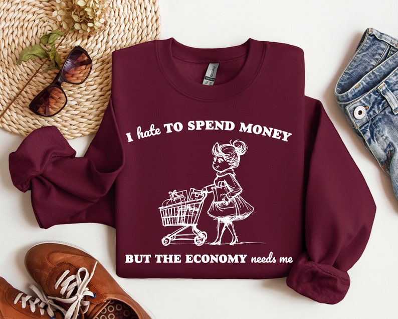 May include: Burgundy sweatshirt with white text that reads "I hate to spend money but the economy needs me". A cartoon illustration of a girl pushing a shopping cart is above the text.